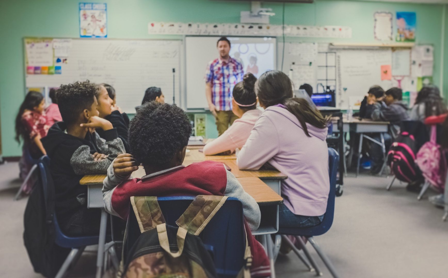 Photograph of adolescent students seated at a table in a school setting, viewed from the back of the classroom, with a teacher in the front of the room